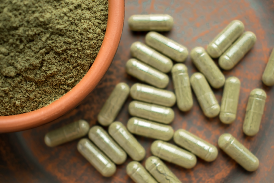 A bird's eye view of kratom powder (in focus) and a lot of kratom capsules (out of focus).