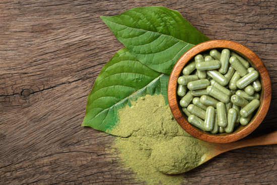 Hulu Kapuas kratom capsules in a bowl next to some kratom powder and kratom leaves.